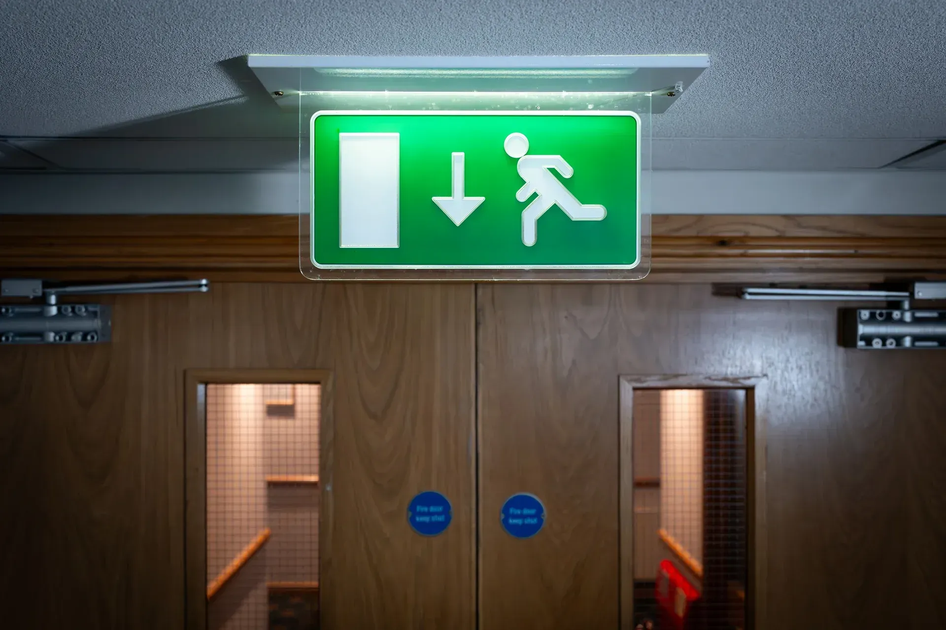 Green illuminated emergency exit sign above double wooden doors in a hallway.