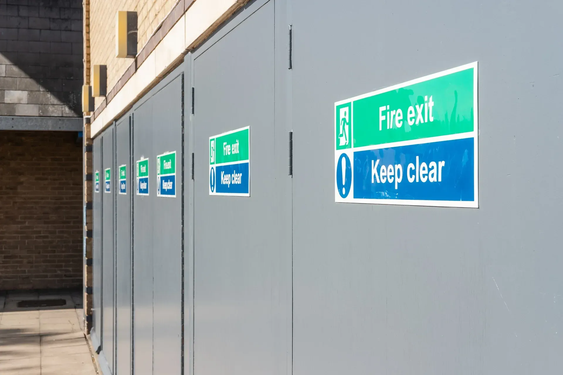 Row of green and blue “Fire exit Keep clear” signs on gray wall in an industrial corridor