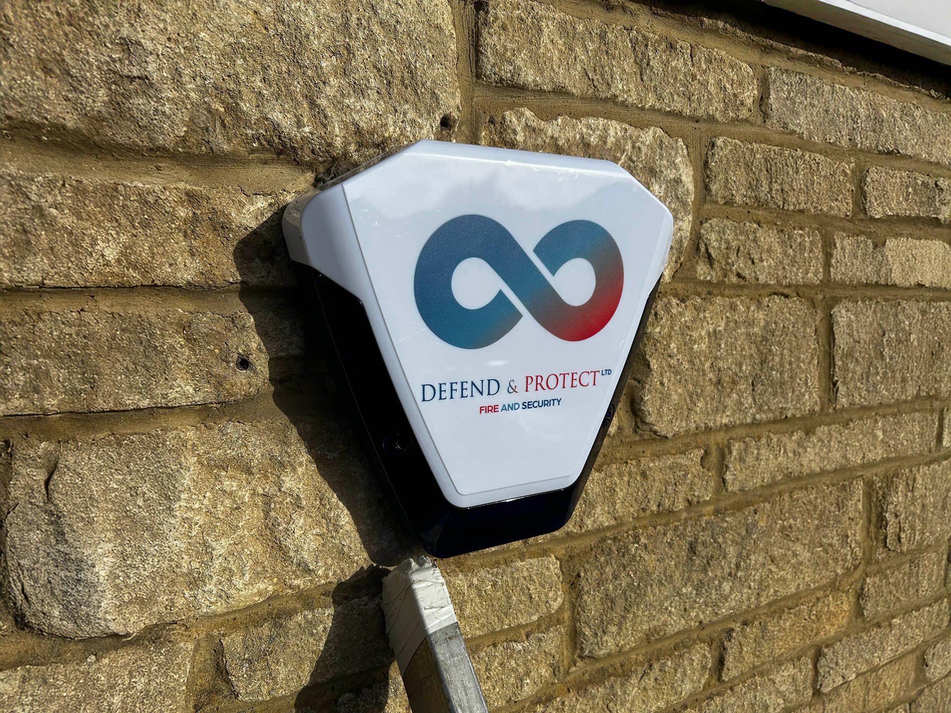 Wall-mounted security alarm box with blue-red logo and "DEFEND PUNCE" text on a stone wall