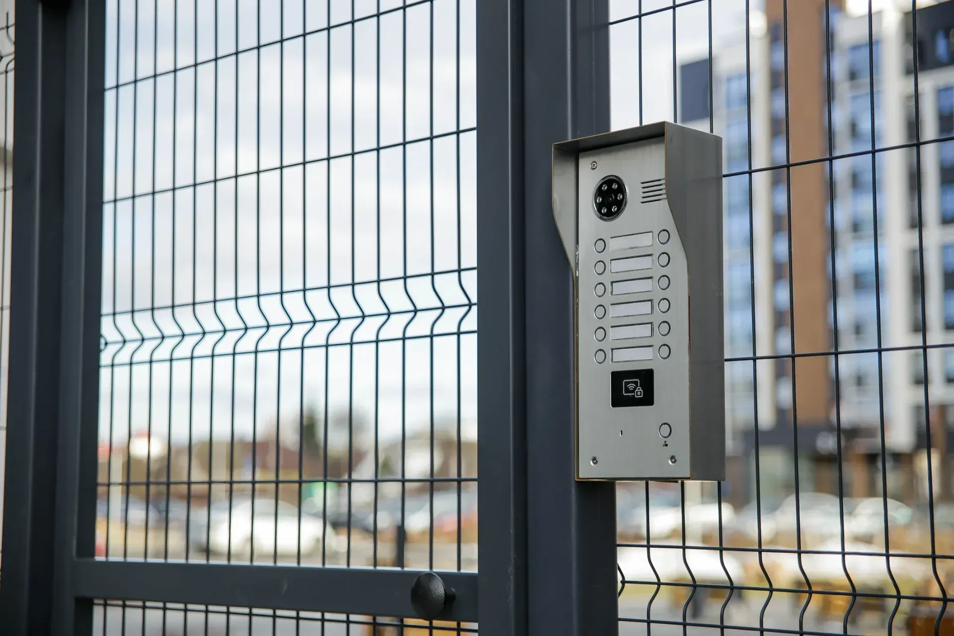 Intercom panel mounted on a metal fence with blurred buildings in the background