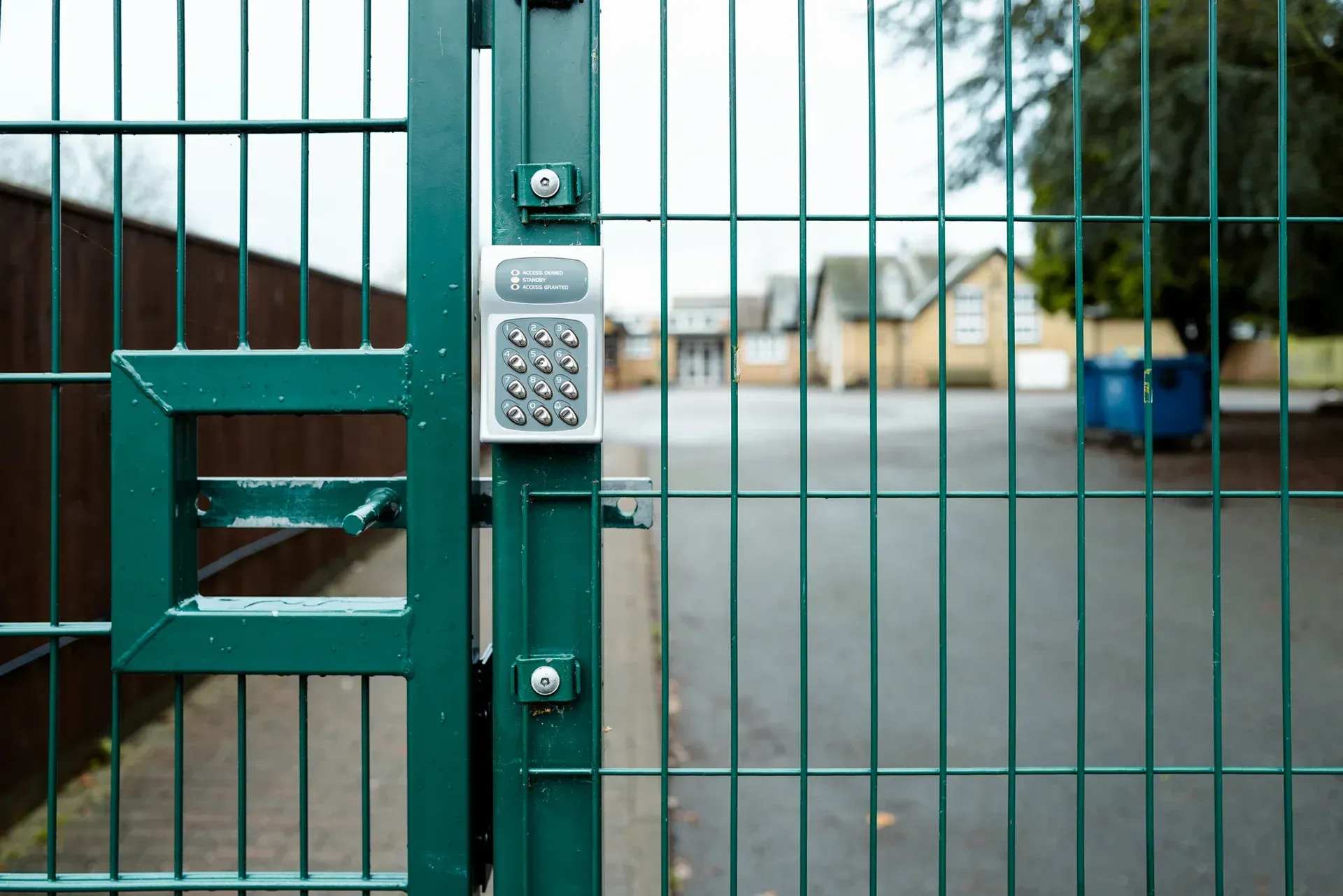 Green metal gate with keypad lock, overlooking a driveway and blurred houses beyond