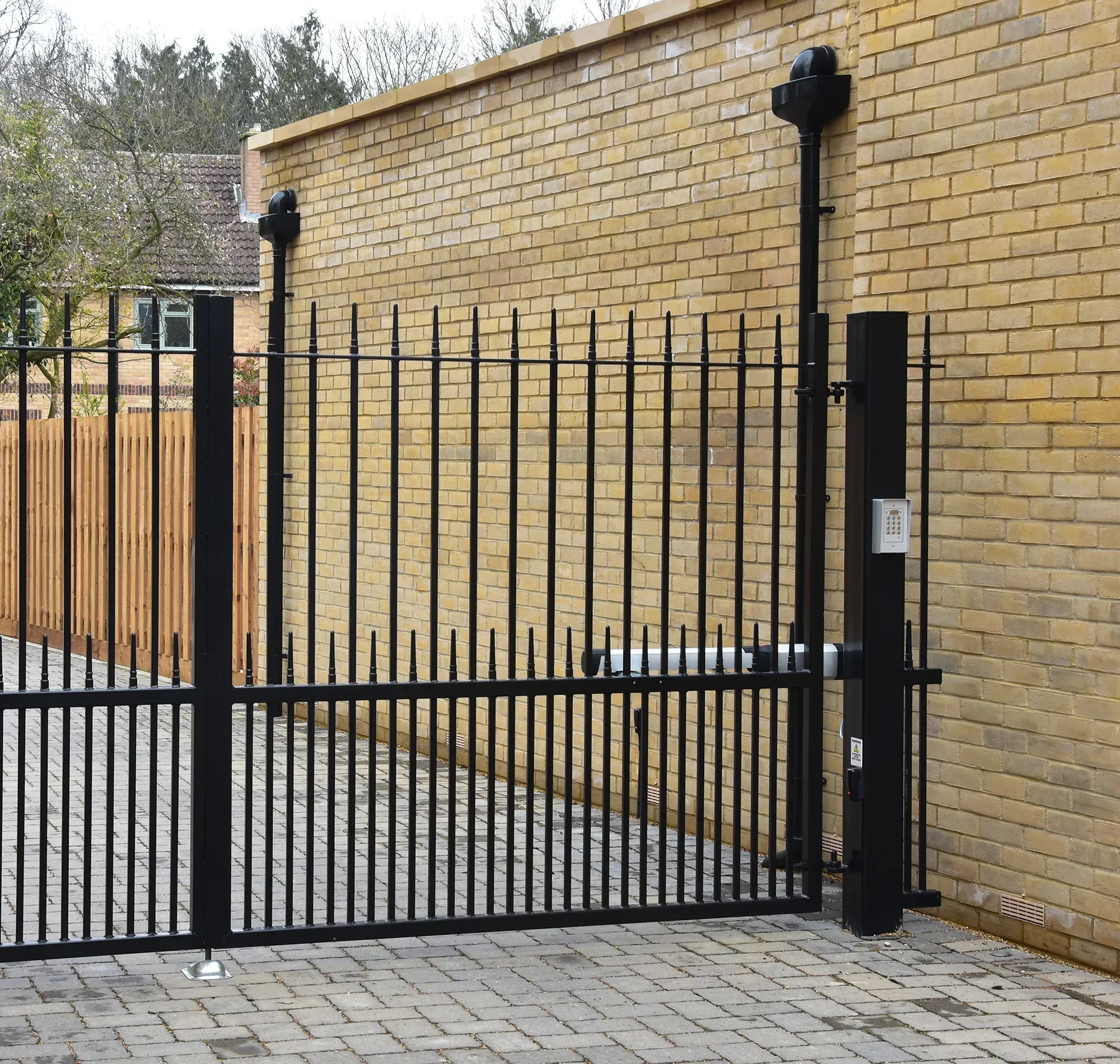 Black metal sliding gate set in front of a yellow brick wall and paved driveway.