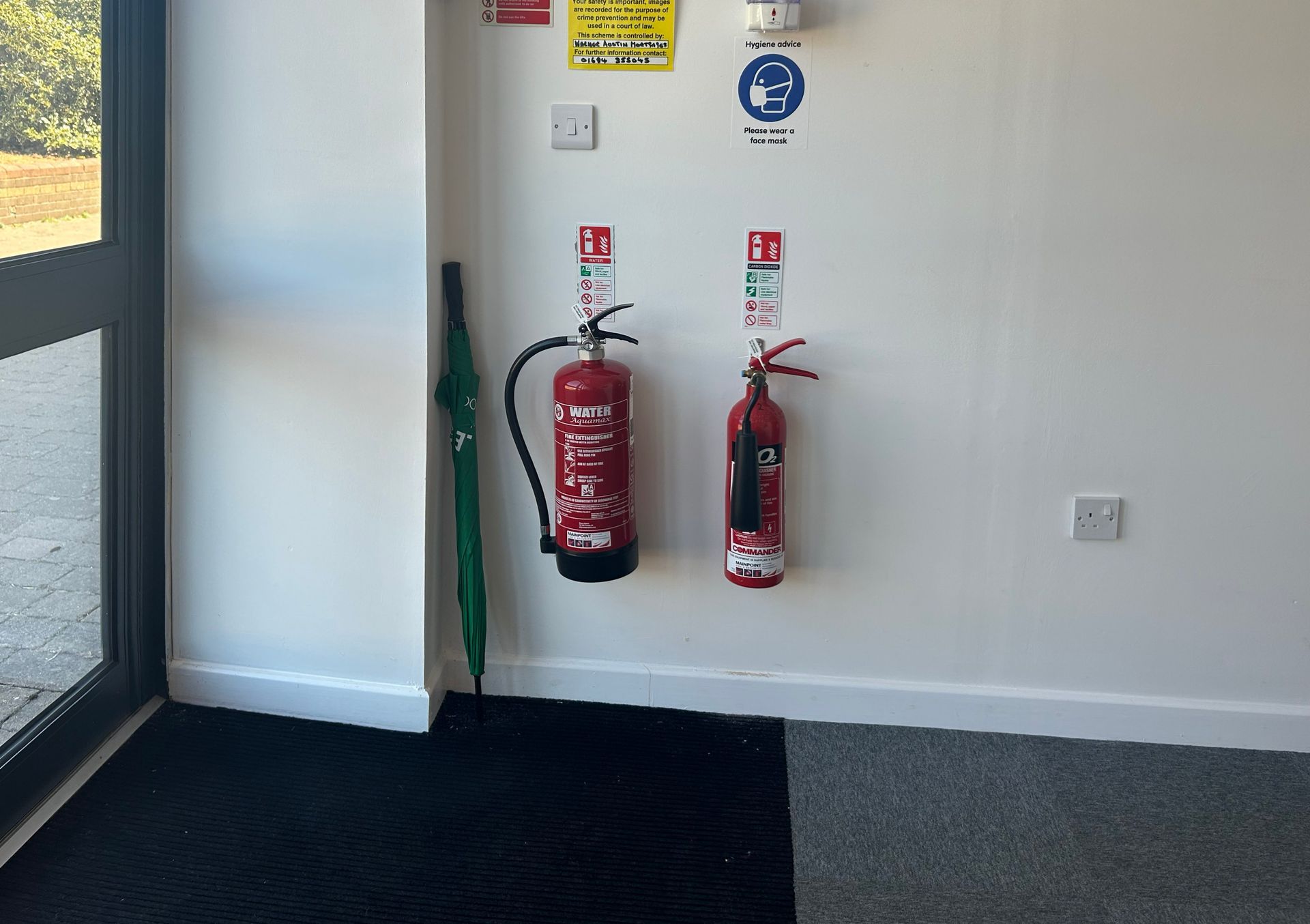Two red fire extinguishers mounted on a white wall beside a green mop and a doorway