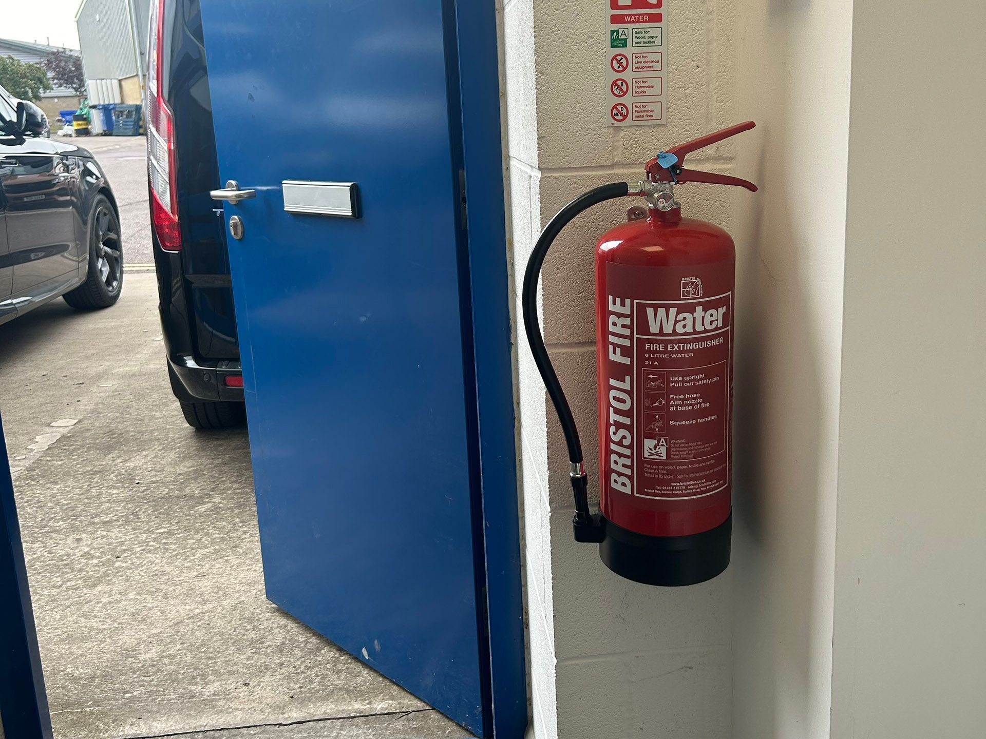 Red fire extinguisher mounted on a wall beside a blue door and parked cars outside.