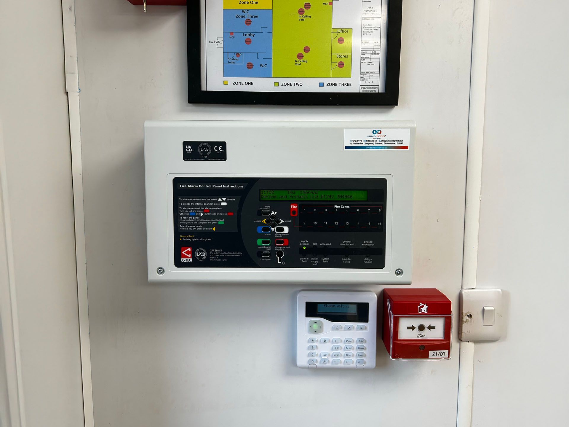 Wall-mounted fire alarm control panel with a small display and red alarm box in a hallway