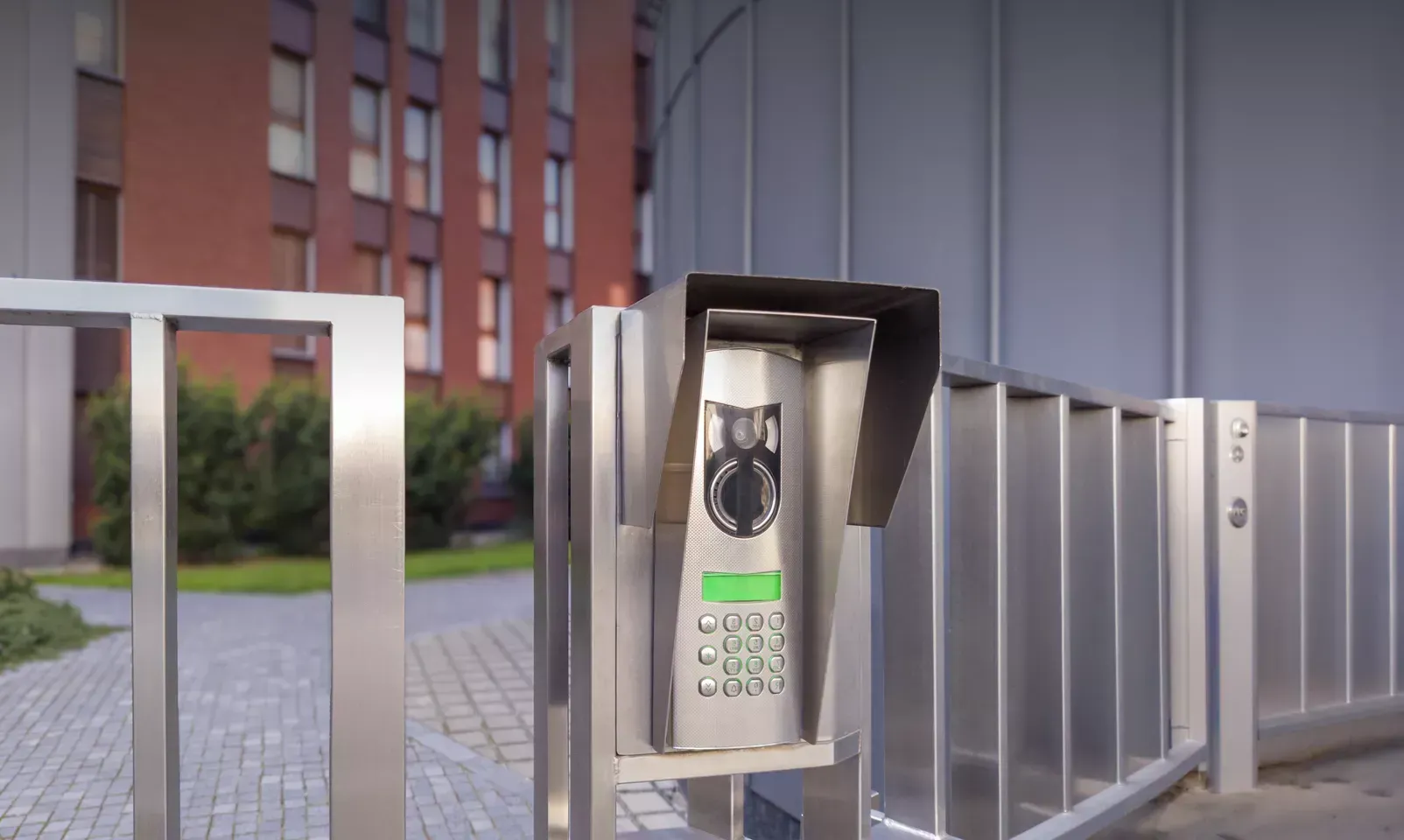 Video intercom keypad at a gated entrance beside a metal fence and brick building
