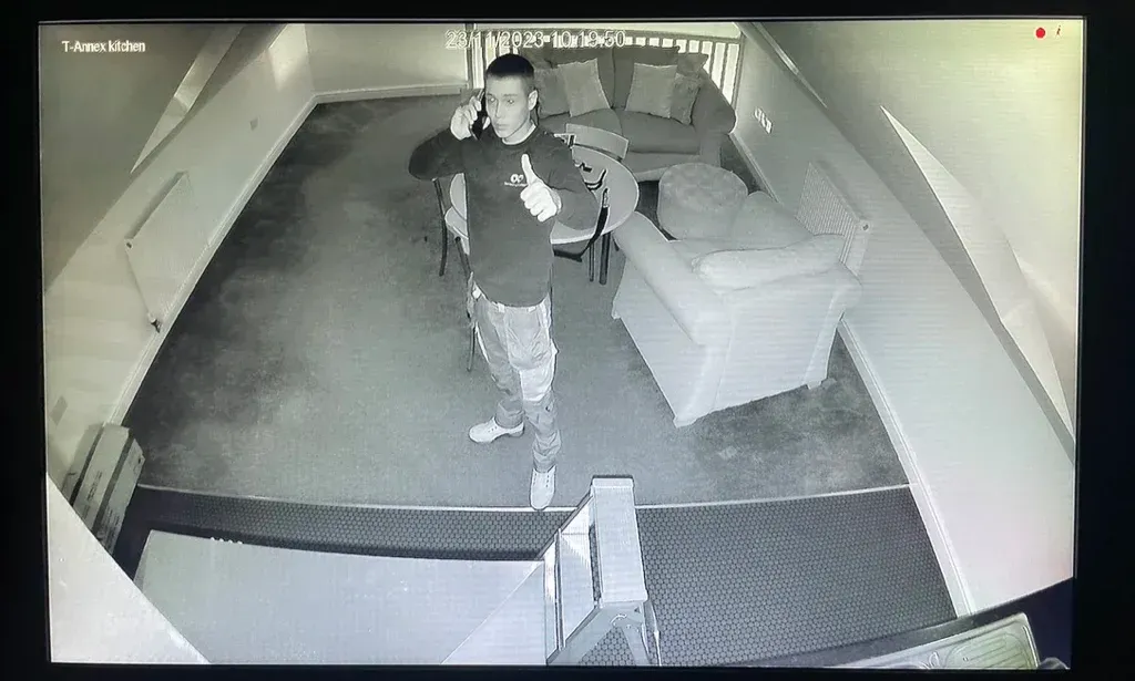 Person walking through a security-camera view of a dim indoor hallway with desks and carpet.