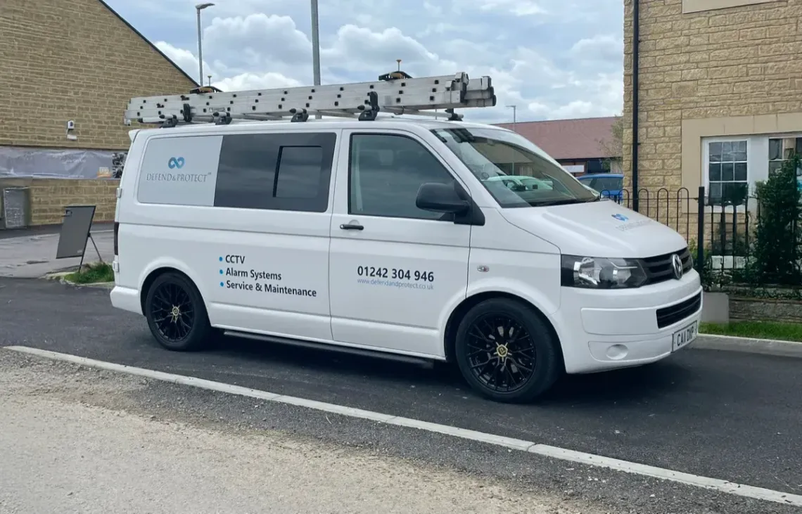 White utility van parked on a street beside a stone building, with ladders on its roof.
