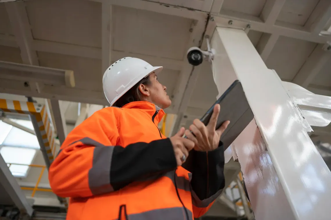 Worker in orange safety jacket and hard hat inspecting a structure with a tablet indoors