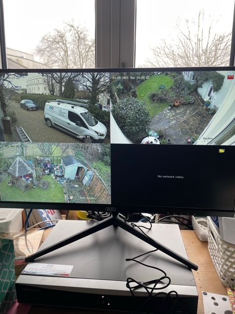 Four-monitor security camera setup on a desk, showing driveway, yard, and patio views.