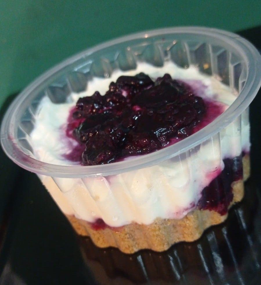 Vaso de plástico transparente con postre en capas cubierto con salsa de frutos rojos oscuros sobre una bandeja negra.