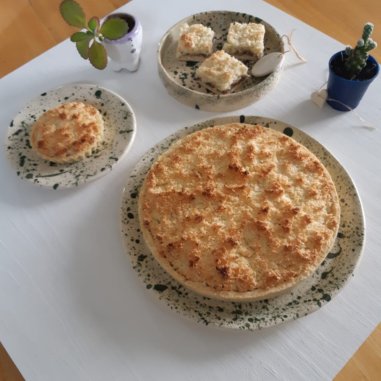 Tres pasteles emplatados sobre un mantel blanco con pequeñas plantas en macetas cerca.