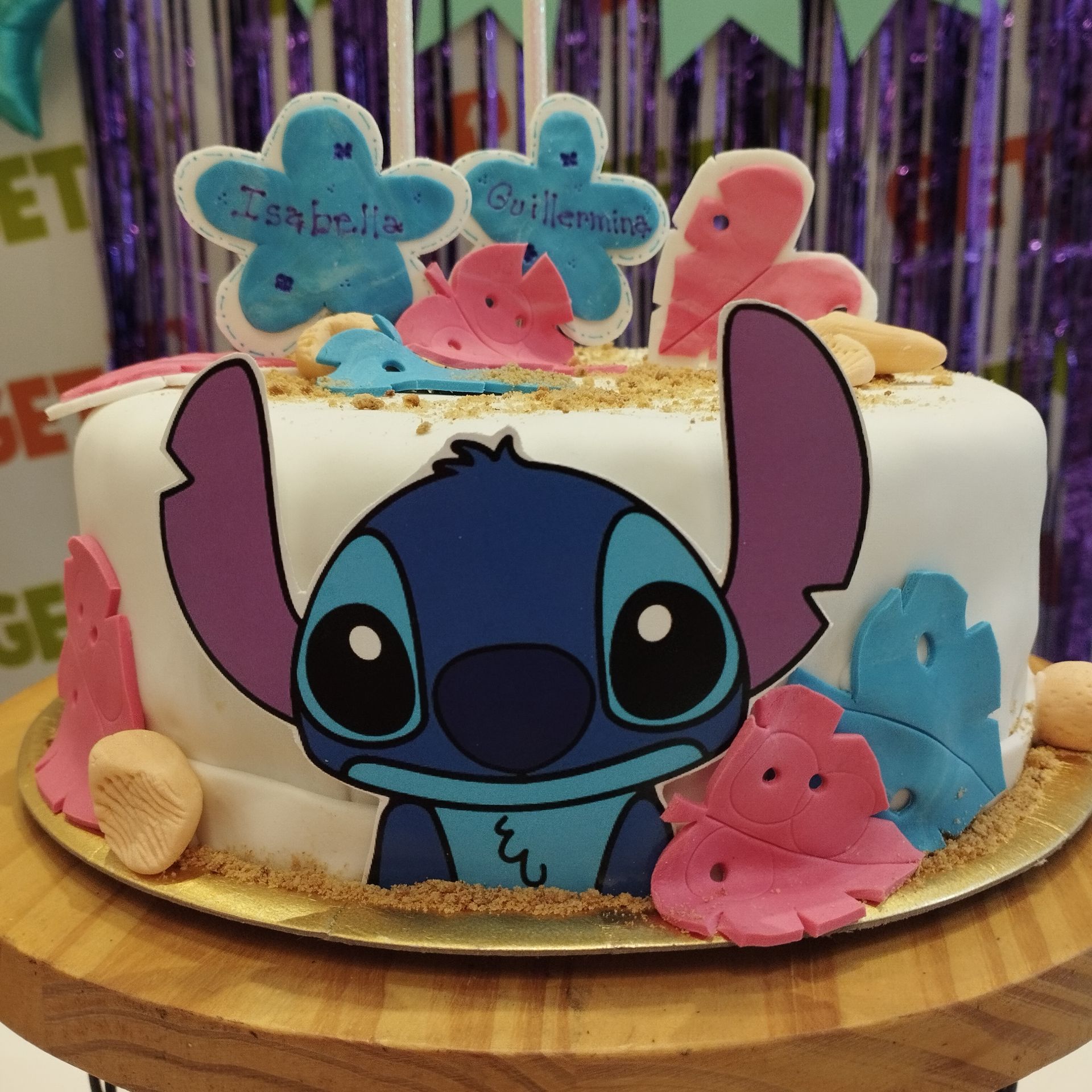 Tarta blanca con temática de Stitch, decorada con motivos azules y rosas sobre una base dorada.