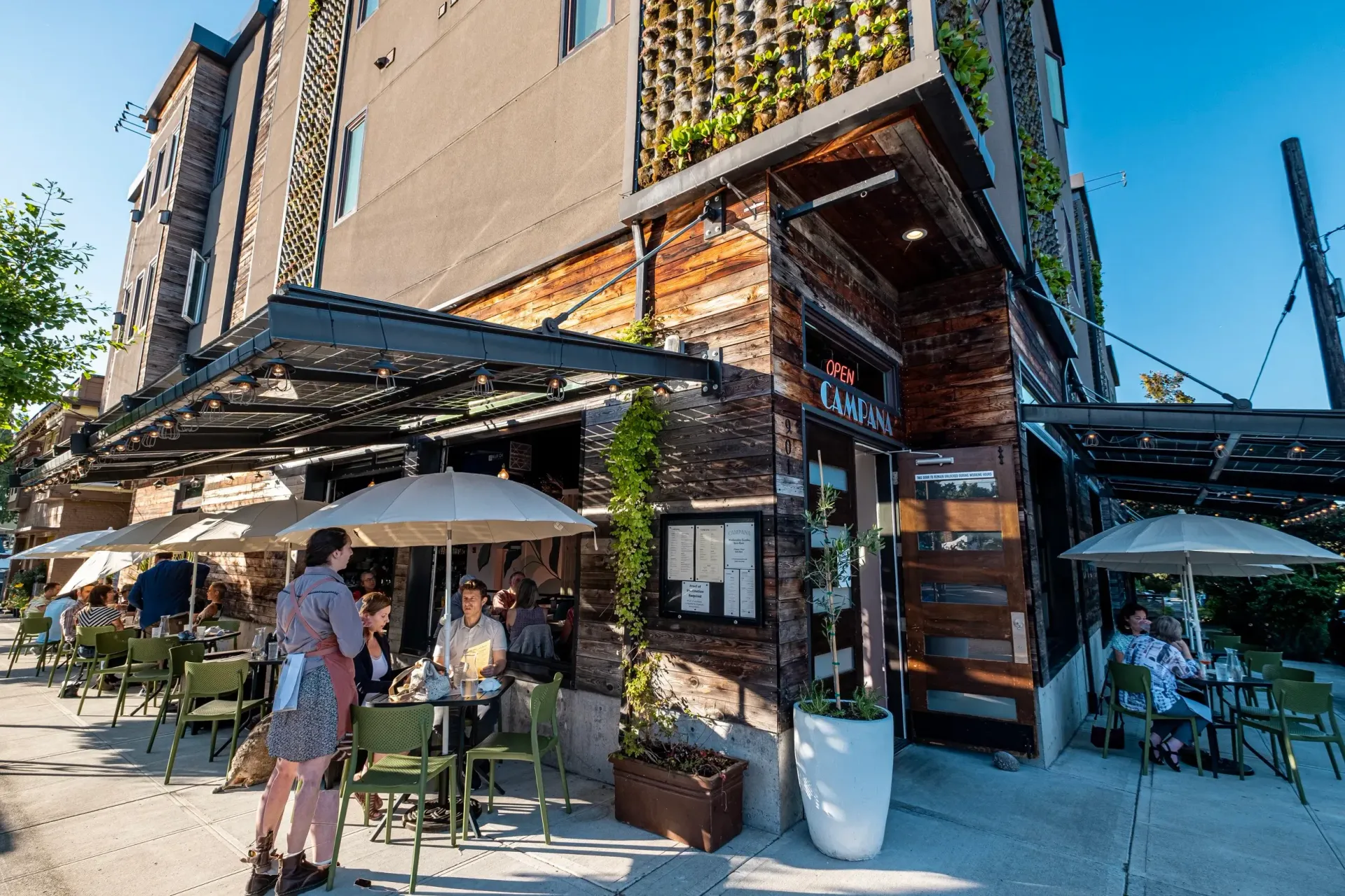 Outdoor café on a sunny street corner with umbrellas, tables, and brick-and-metal storefront seating