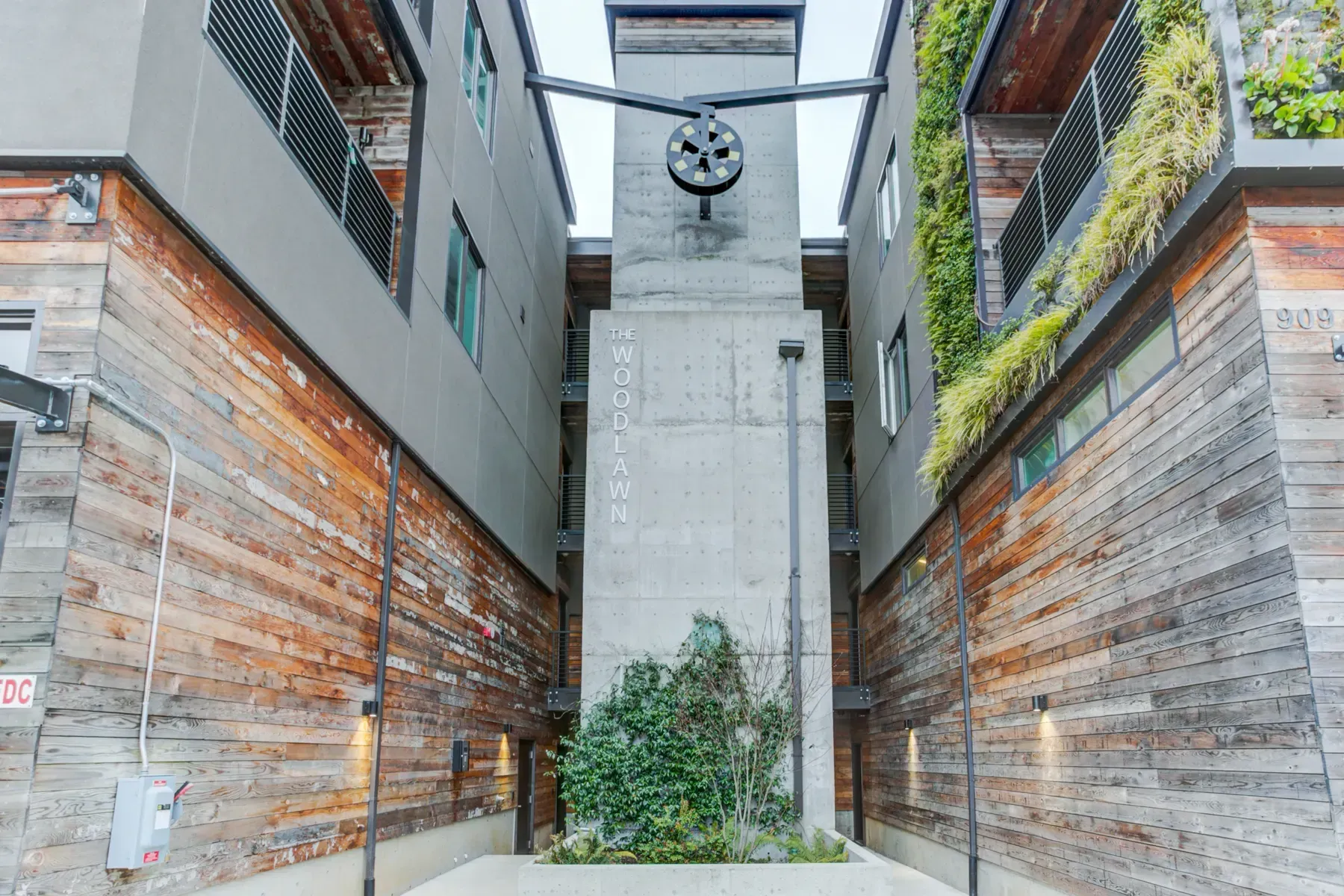 Narrow courtyard with modern buildings, wood and brick walls, and a small central tree beneath a clock-like fixture