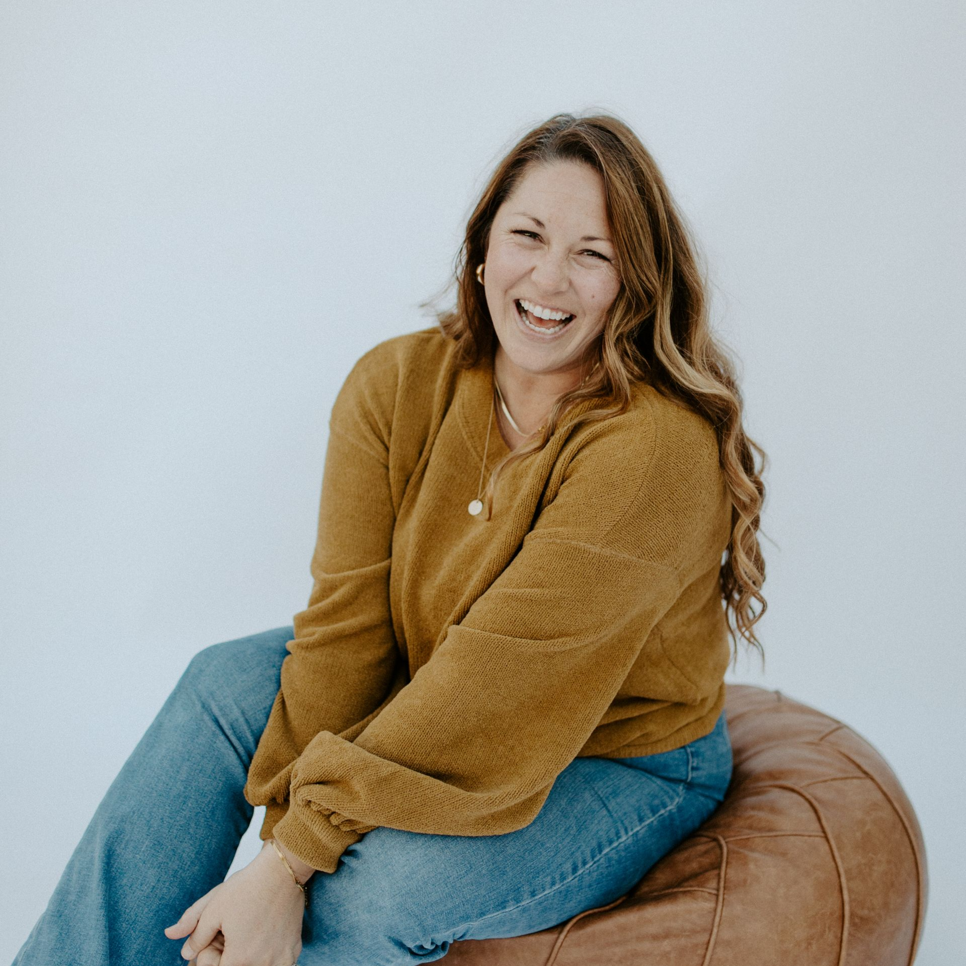 Dr. Steph - Smiling woman in mustard sweater and jeans sitting on a brown pouf against a white wall