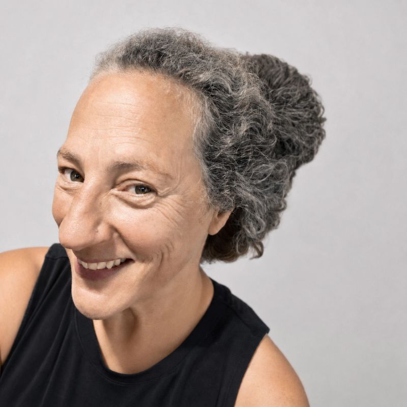 Dinnerman - Smiling person with gray curly hair in a black sleeveless top against a light background