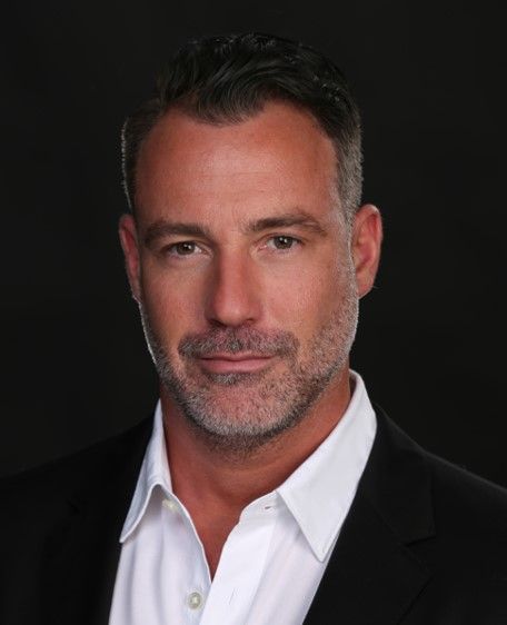 Dr. Brian Capra - Studio headshot of a man in a black suit jacket and white shirt against a dark background