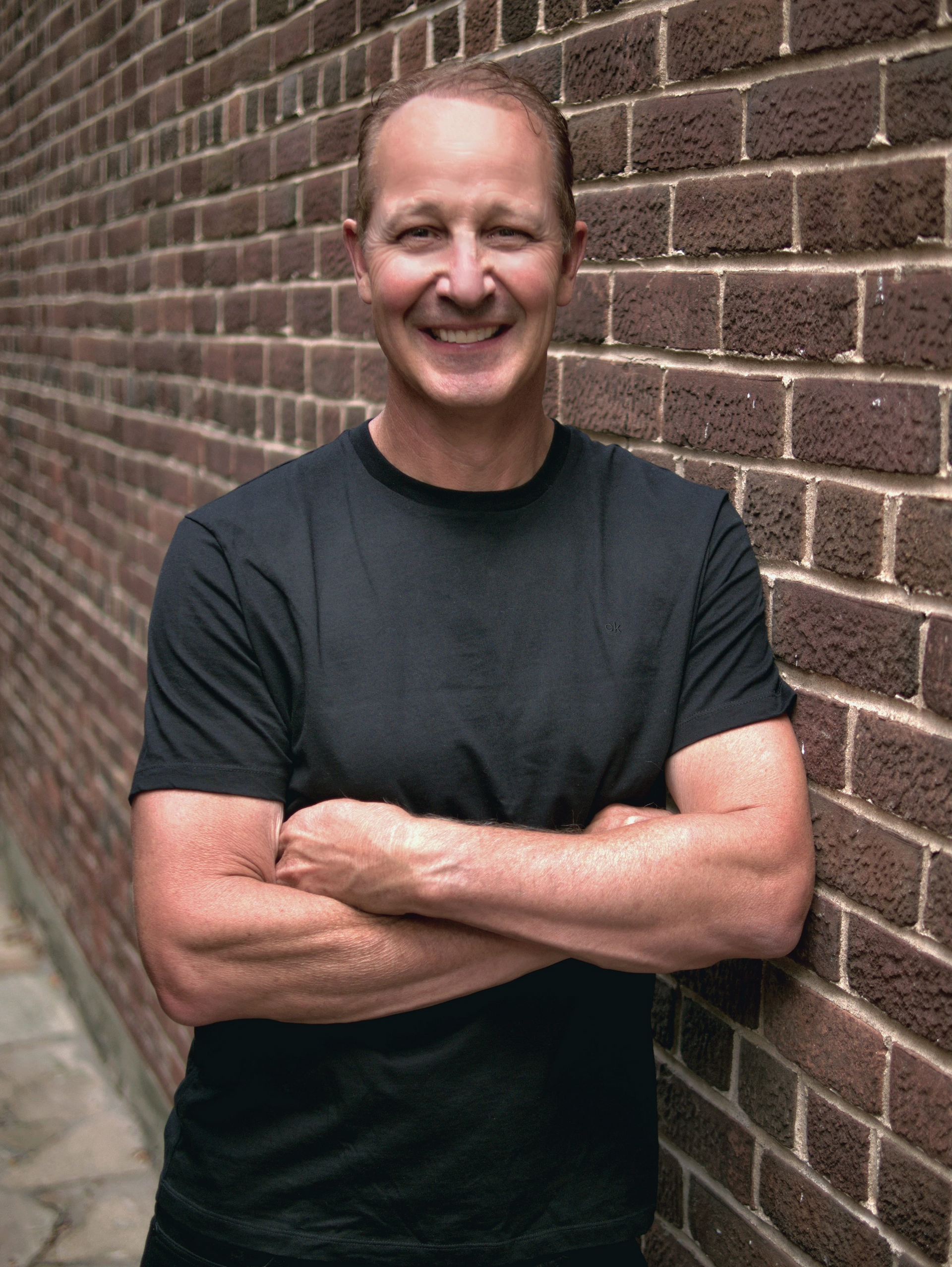 Dr David Fletcher - Smiling man in a black T-shirt standing with arms crossed against a brick wall