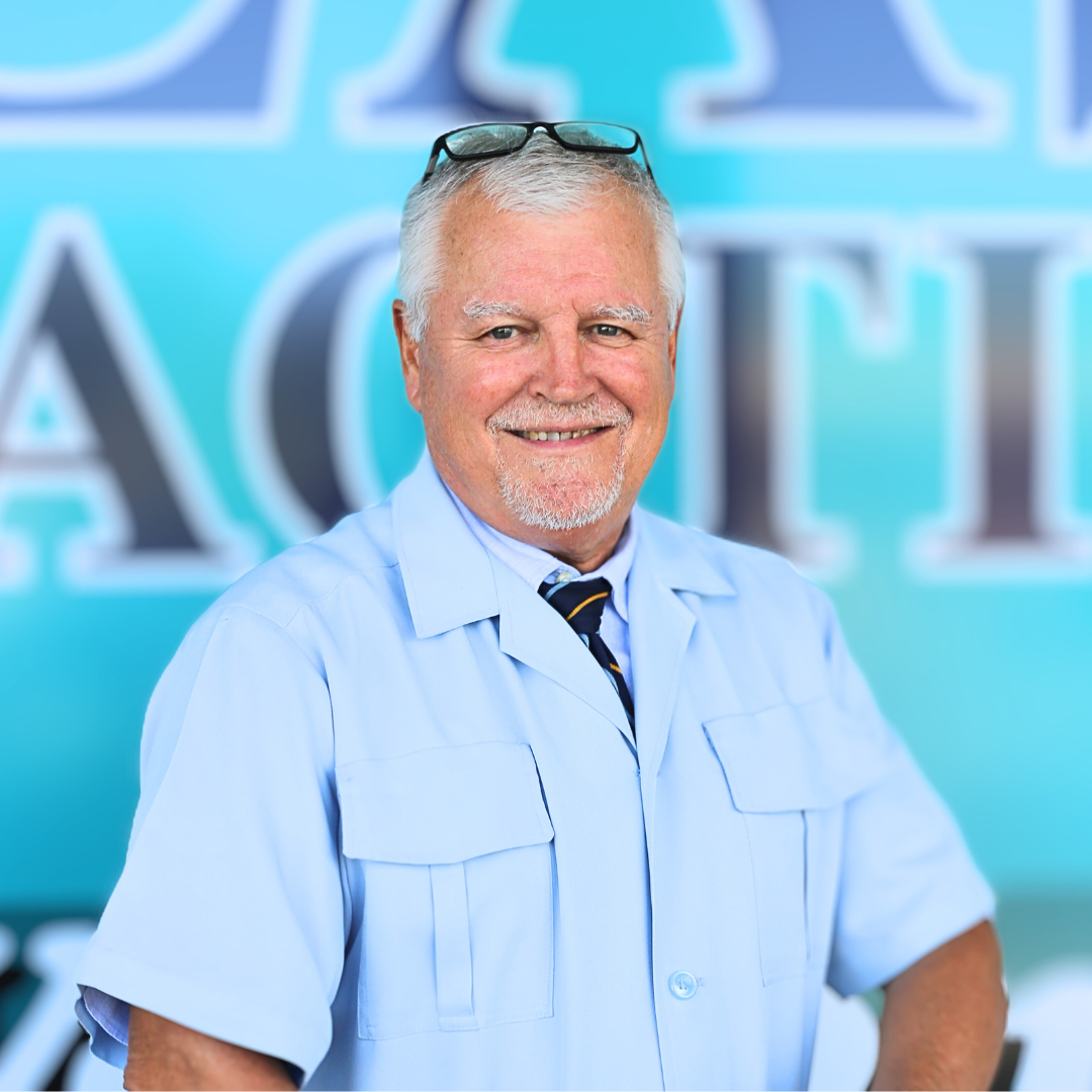 Dr. Fred - Smiling man in a light blue shirt and glasses stands before a blurred blue sign