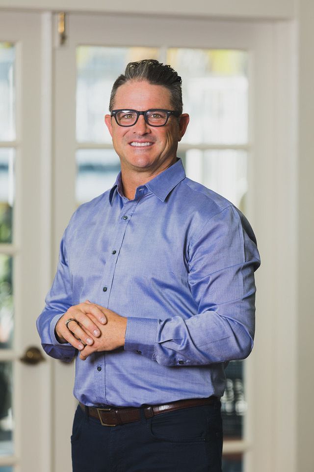 Dr. Matt Hubbard - Man in a blue shirt and glasses standing indoors, smiling with hands clasped.