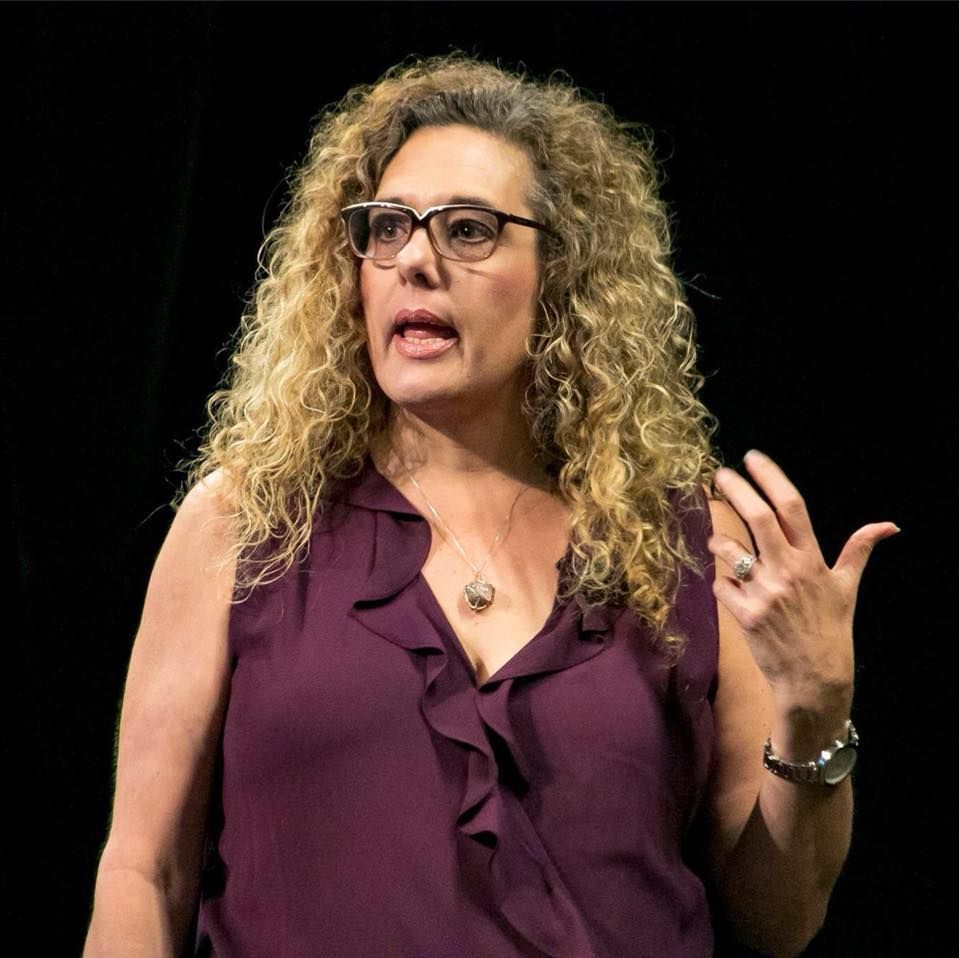 Dr. Monika Buerger - Woman with curly blond hair speaking on stage, wearing glasses and a purple sleeveless blouse