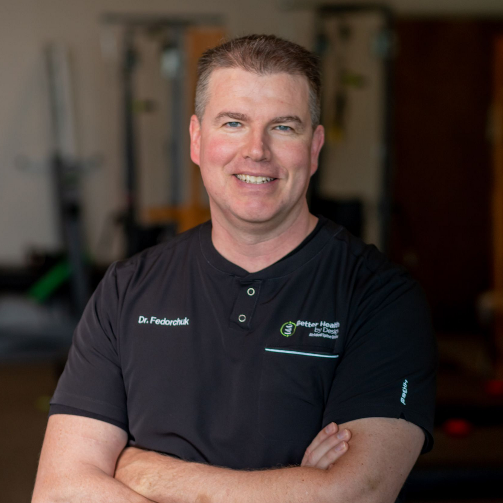Dr Curtis Fedorchuk - Smiling man in a black polo shirt standing with arms crossed in a gym.