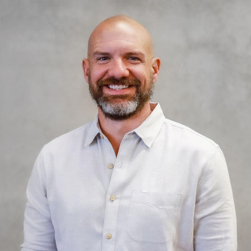 Dr. Tony Ebel - Smiling bald man with a beard in a light gray shirt against a gray background