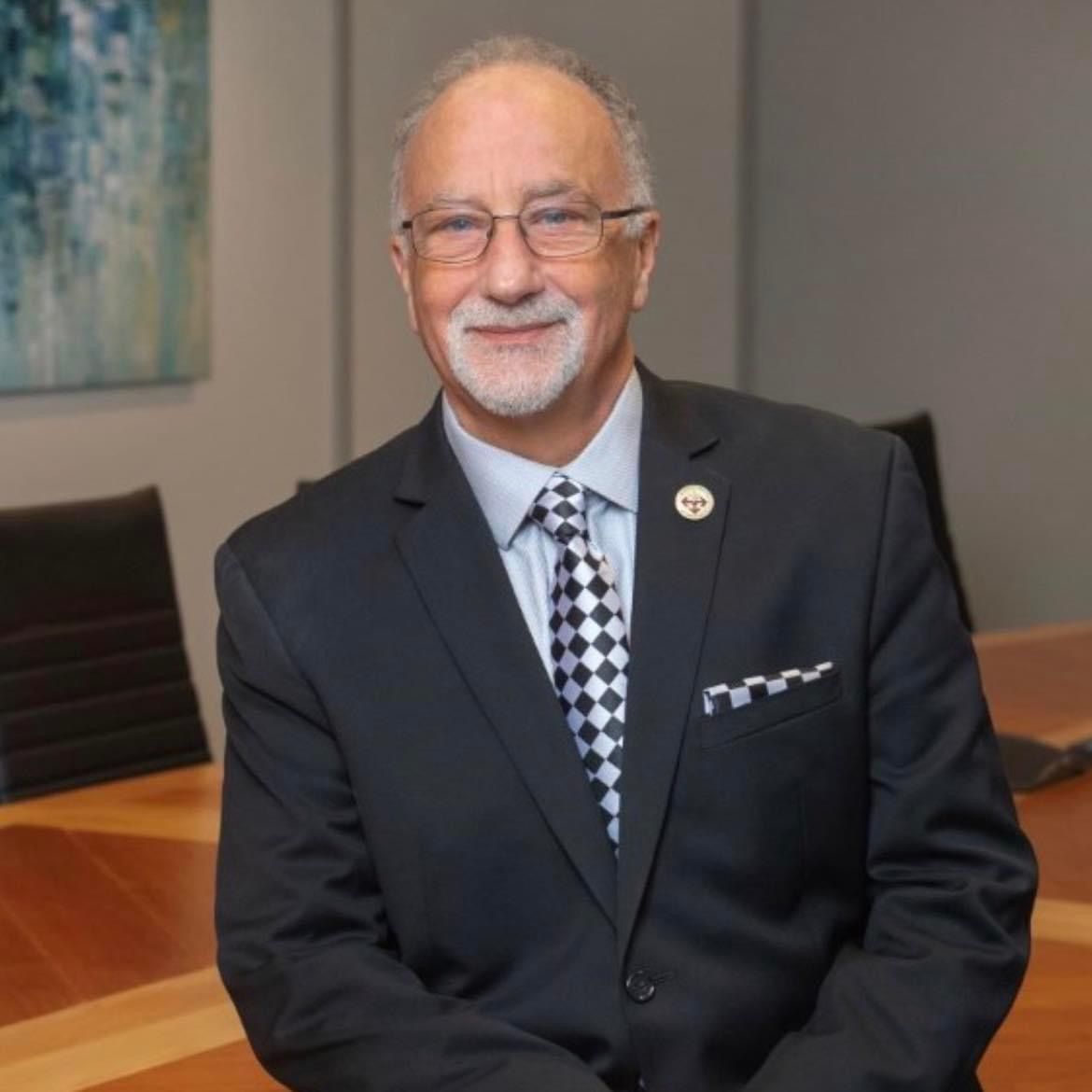 Dr. Jack Bourla - Smiling man in a dark suit seated at a conference table in an office.