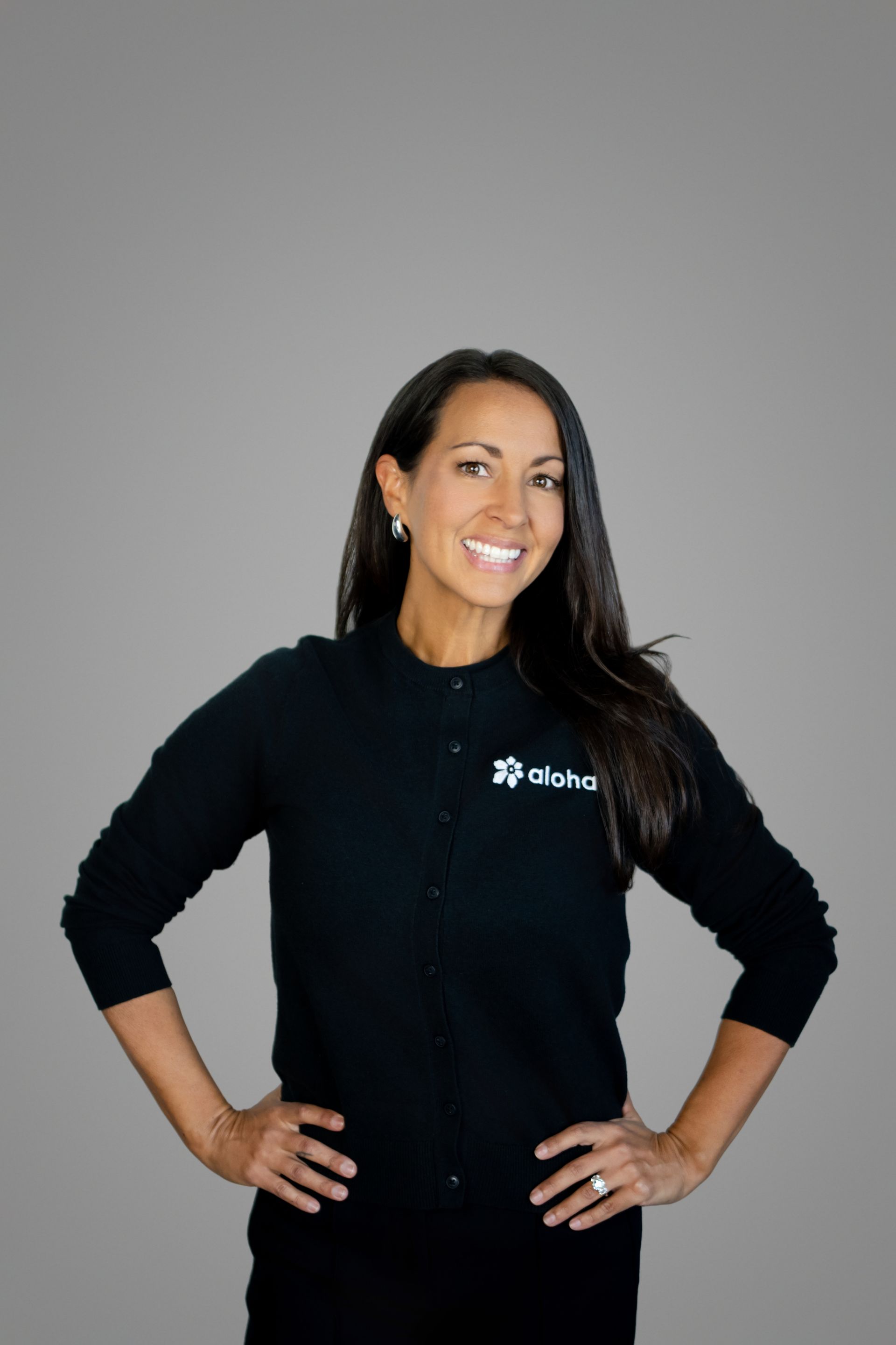 Jenn Denham - Smiling woman in black top, standing with hands on hips against a gray background