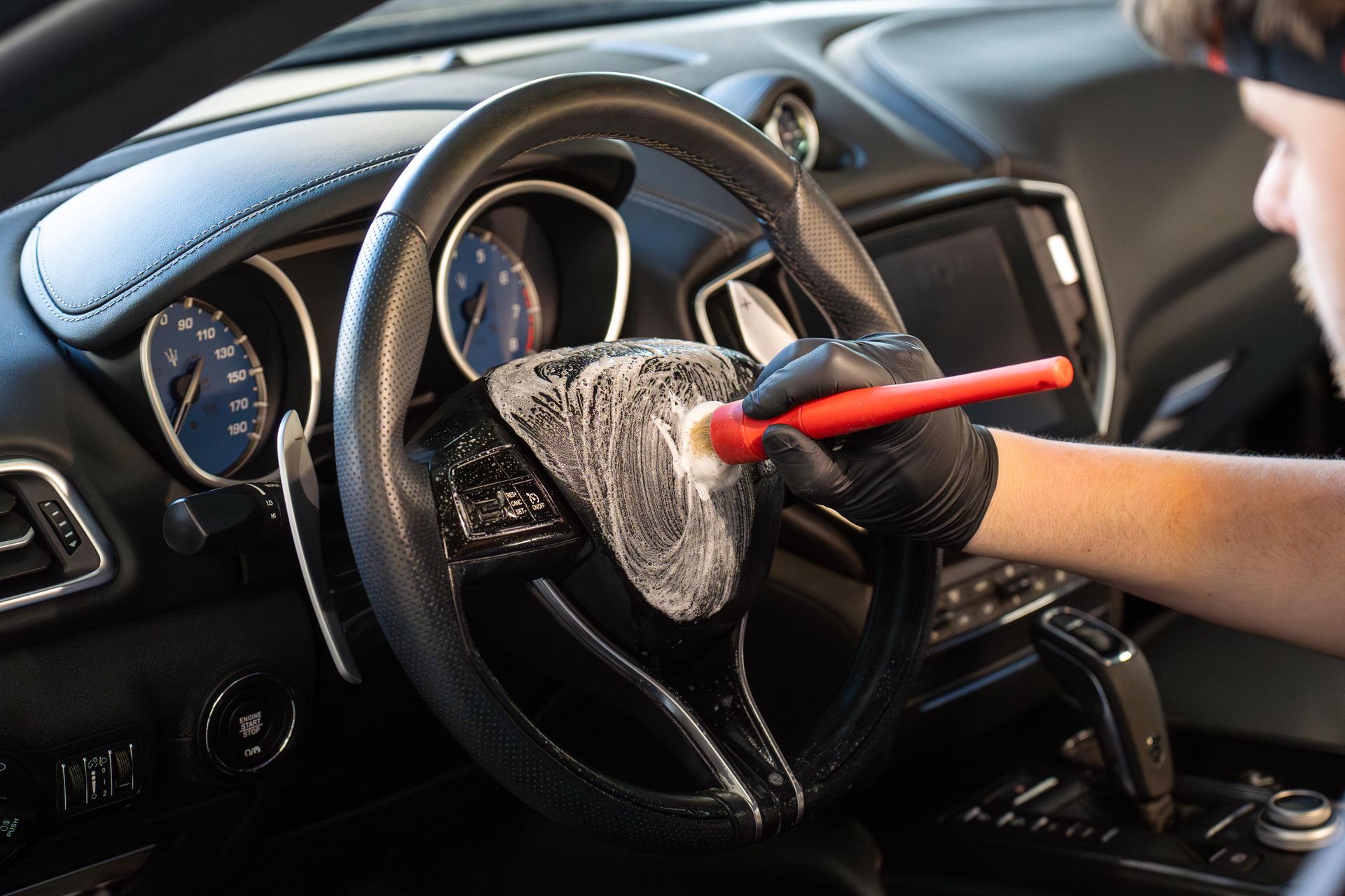 Person cleaning a car's black steering wheel with a red brush and soapy foam. The interior dashboard and gauges are visible.
