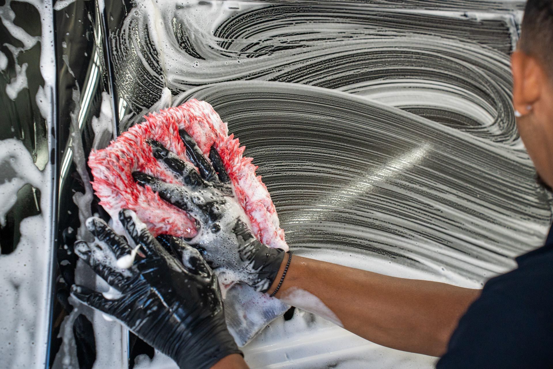 Person wearing black gloves washes a car with a foamy sponge. Soap and water cover the car's surface.