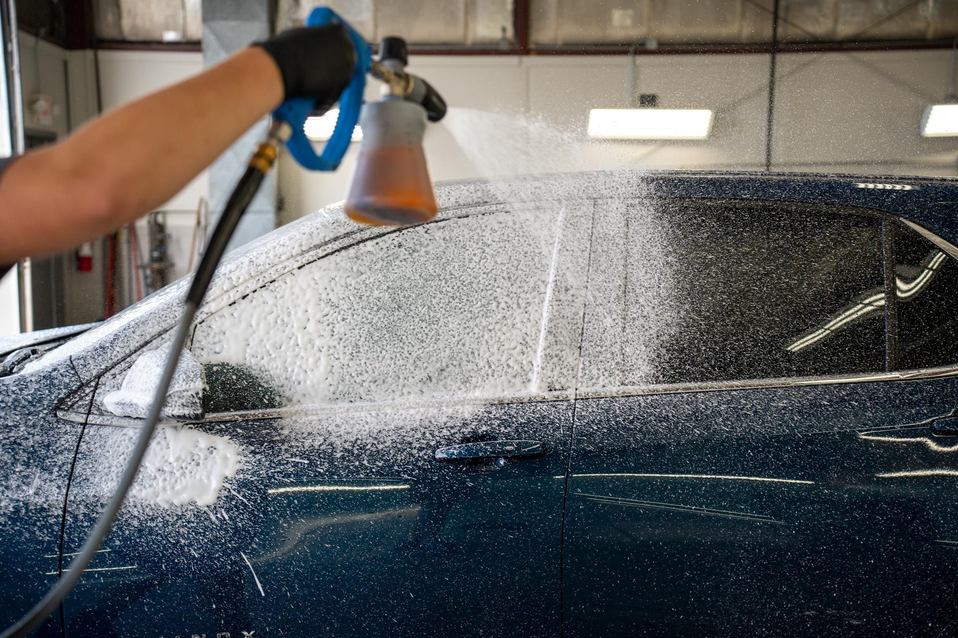 Person spraying soapy foam on a dark blue car, likely for cleaning, in a garage setting.