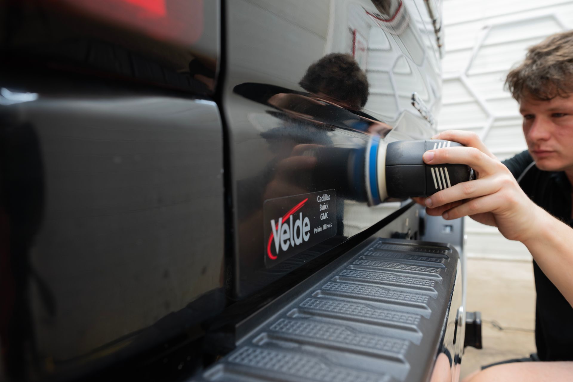 A person polishes a black pickup truck's bumper with a handheld machine outdoors.