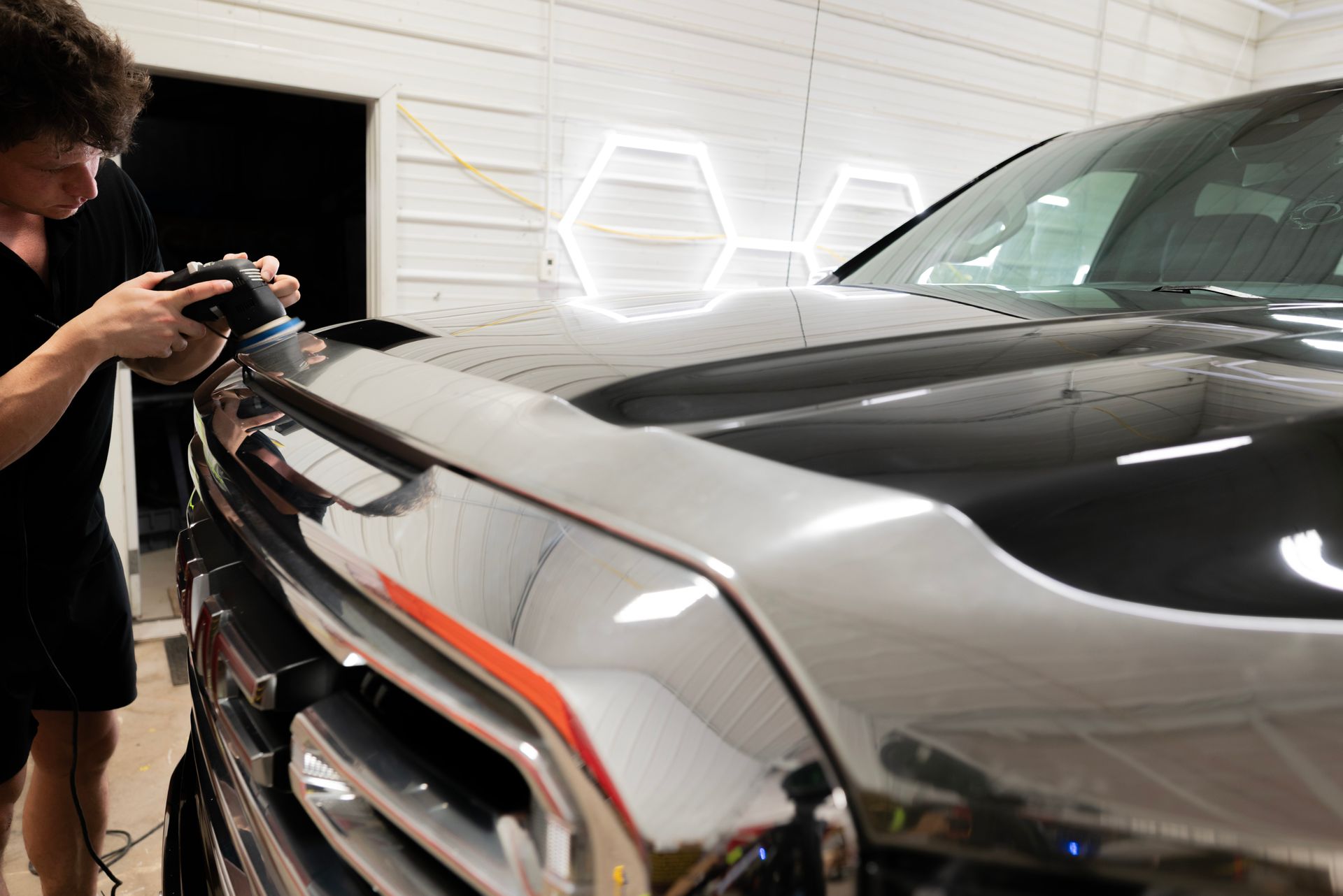 A person polishing the hood of a shiny black pickup truck. The setting is a well-lit garage.