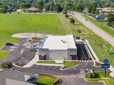 Aerial view of a modern white building with parking lot, road, and green field nearby