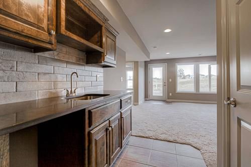 Modern kitchen with dark wood cabinets, tiled backsplash, and open view to a carpeted living area with large windows
