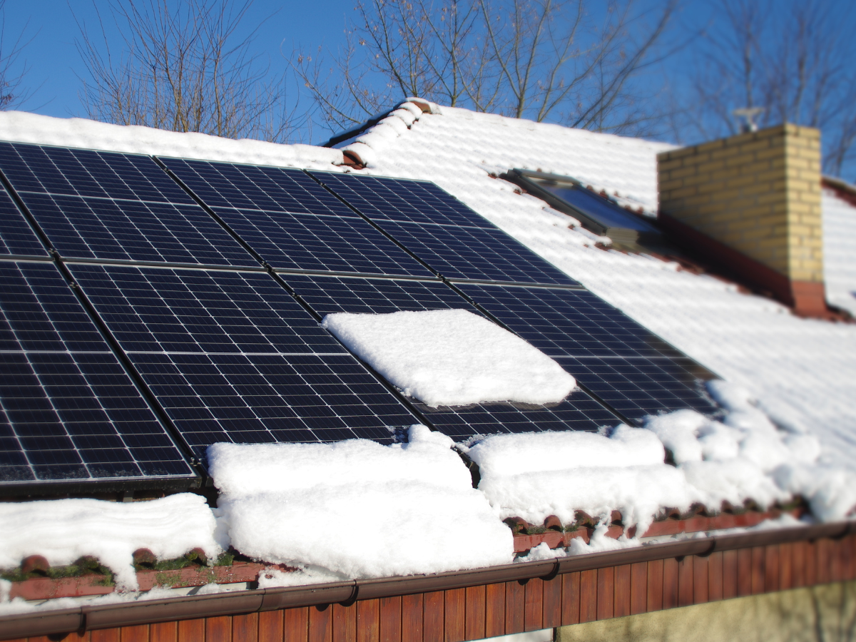 Snow-covered solar panels on a rooftop with a chimney and blue sky