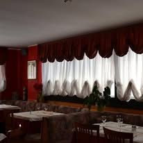 Interno del ristorante con mantovane color bordeaux, tende bianche, tavoli e sedie.