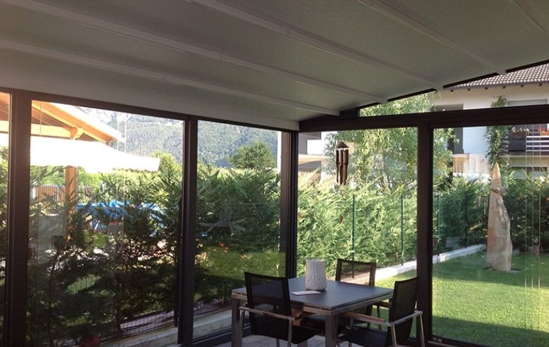 Sunroom with glass walls overlooking a lush green yard, patio furniture, and a retractable roof.