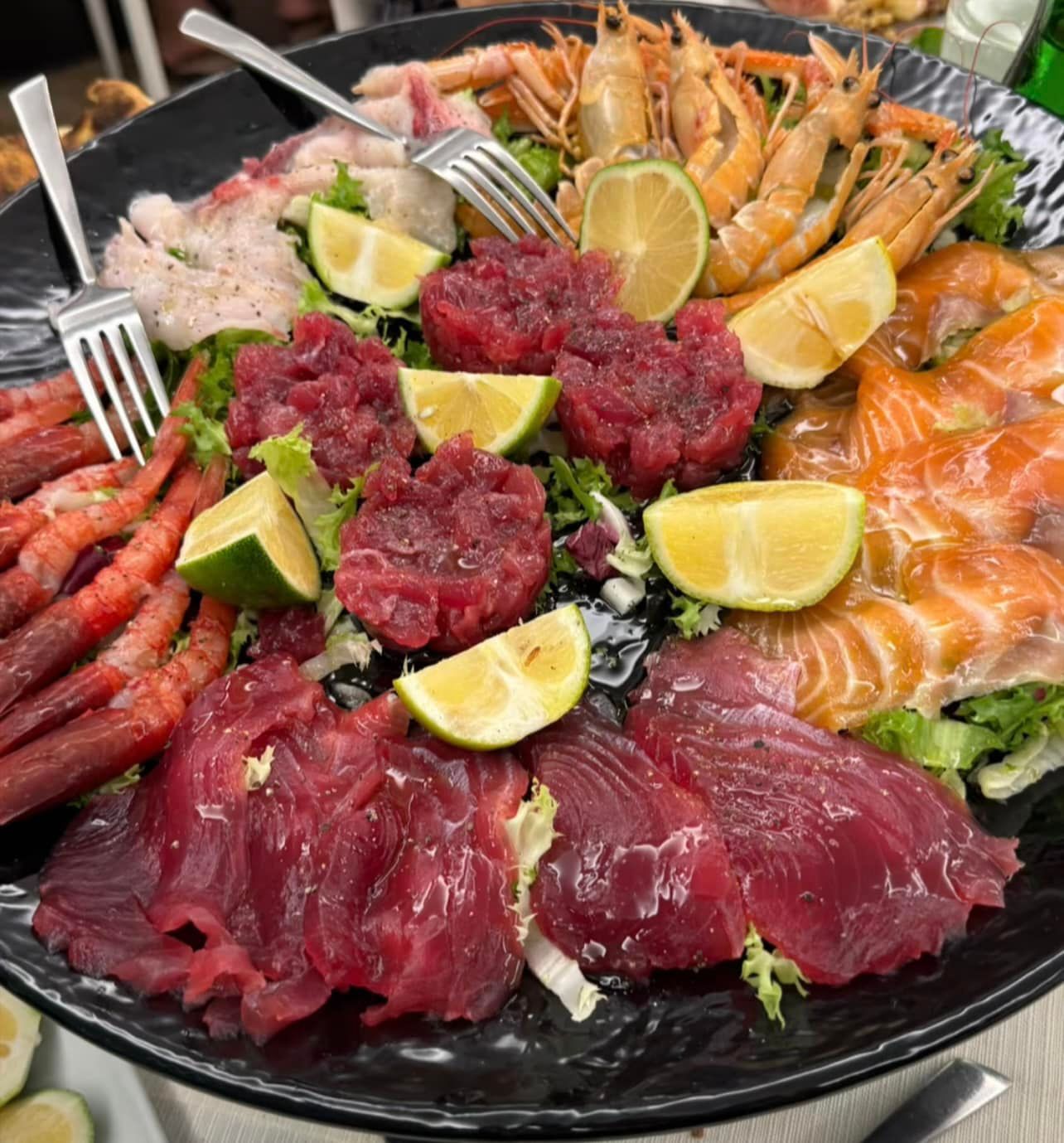 Piatto di frutti di mare: tonno crudo, salmone, gamberi e spicchi di lime su piatto nero.