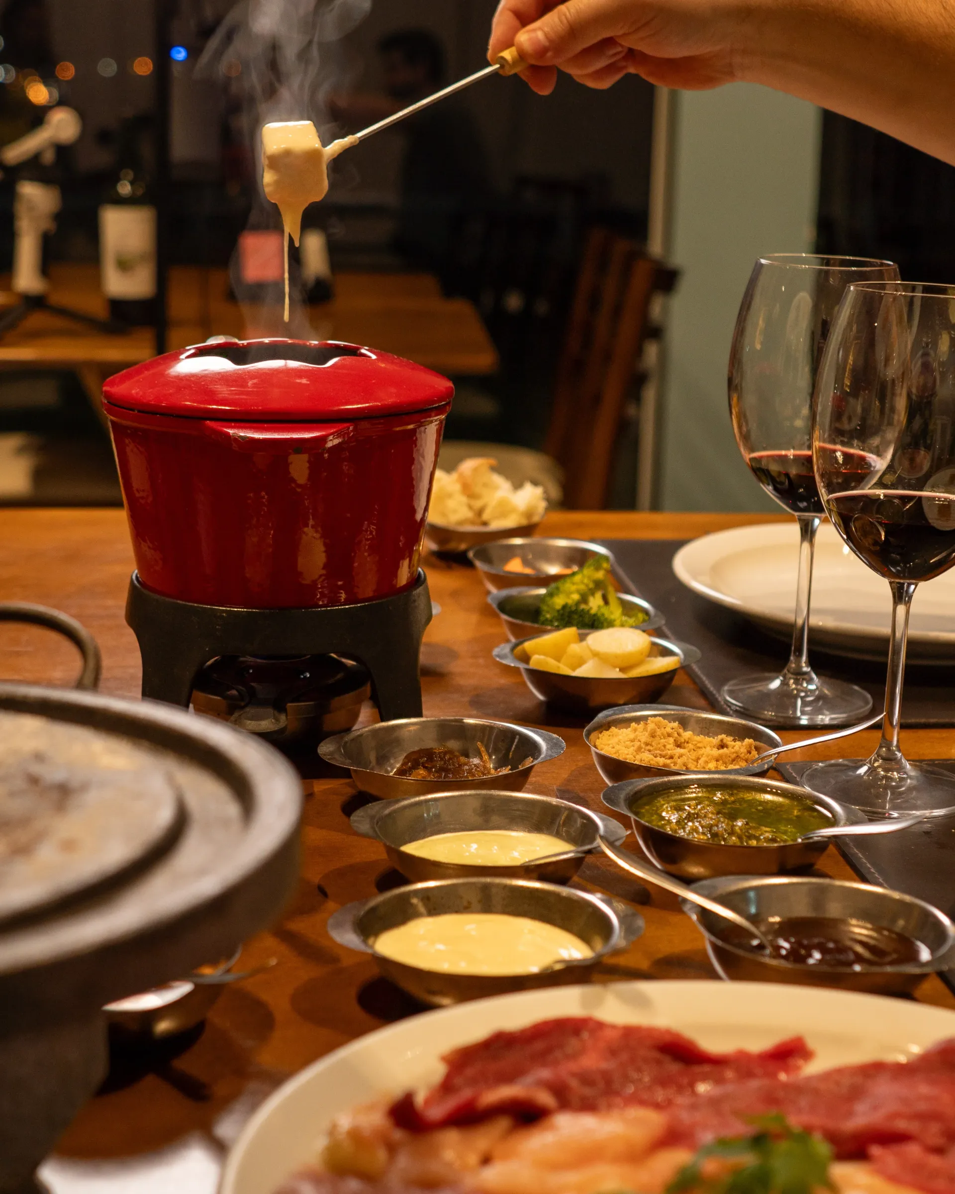 Uma pessoa mergulha comida em uma panela de fondue, com vários molhos e taças de vinho tinto sobre uma mesa. Chalé Mont Hermon.