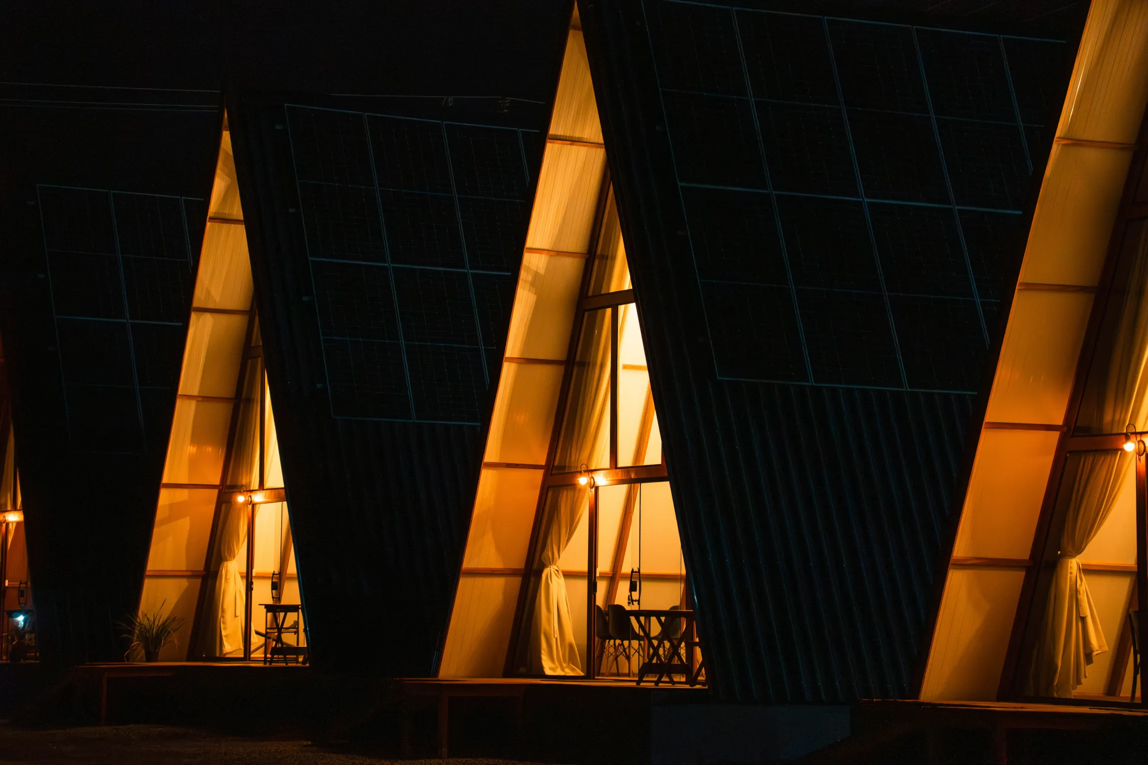 Estruturas triangulares com iluminação interna aconchegante à noite. Mesas e cadeiras visíveis no interior. Chalé Mont Hermon.