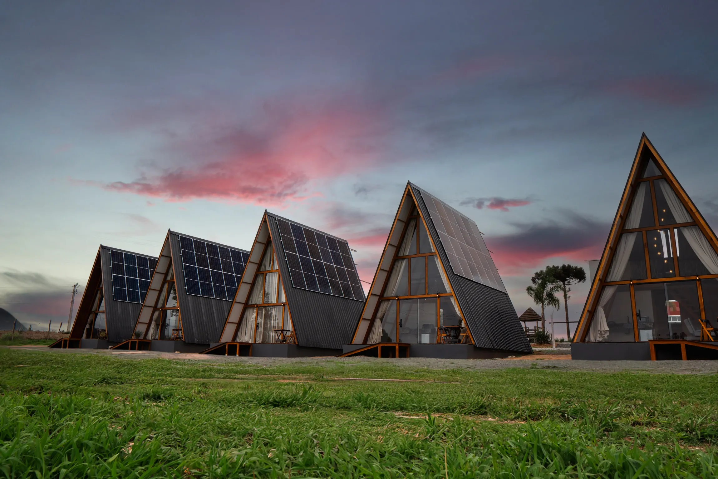 Cabines triangulares com painéis solares contra um céu de pôr do sol sobre um campo gramado.