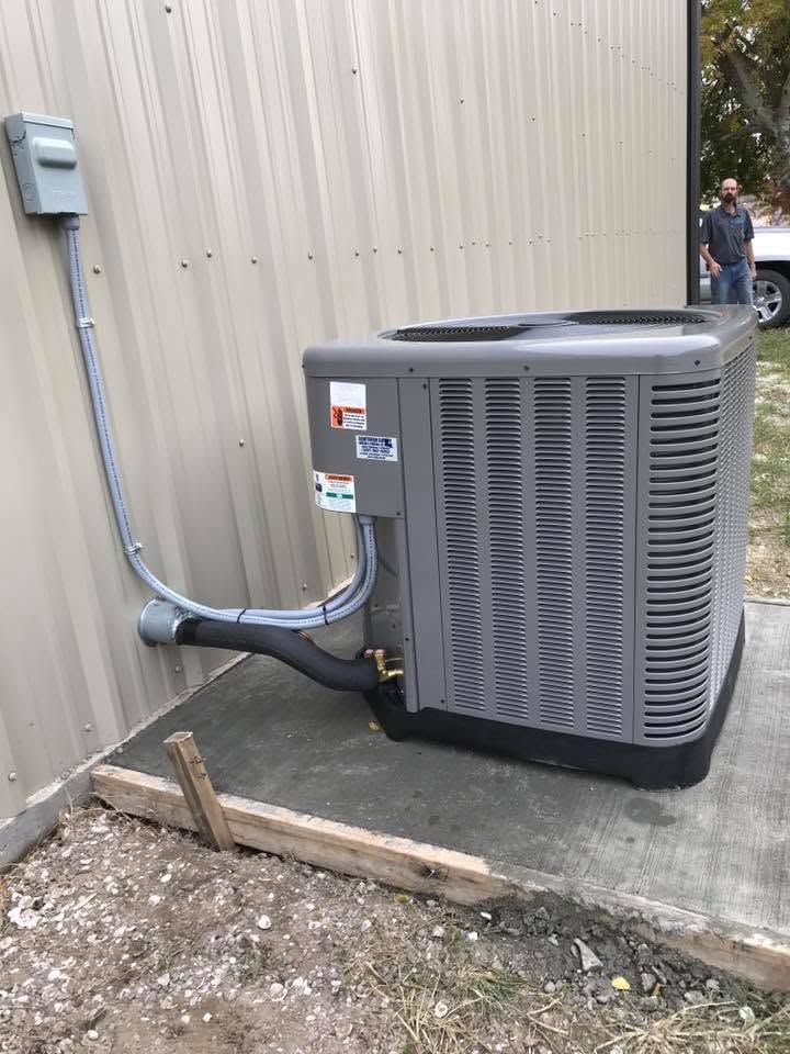 Gray outdoor HVAC unit beside a beige building, with conduit and ducts connected on a concrete pad.