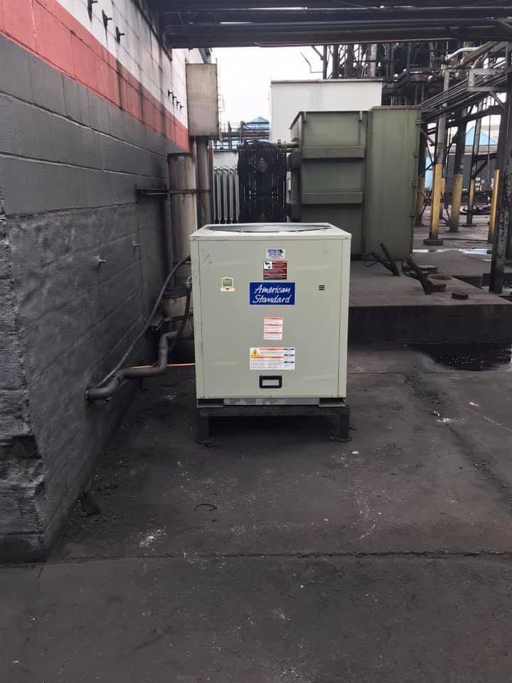 Gray industrial electrical cabinet on a concrete lot beside a warehouse wall and machinery.