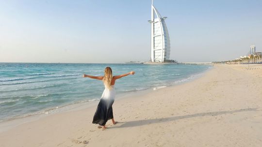 A woman is standing on a beach with her arms outstretched in front of a large building.