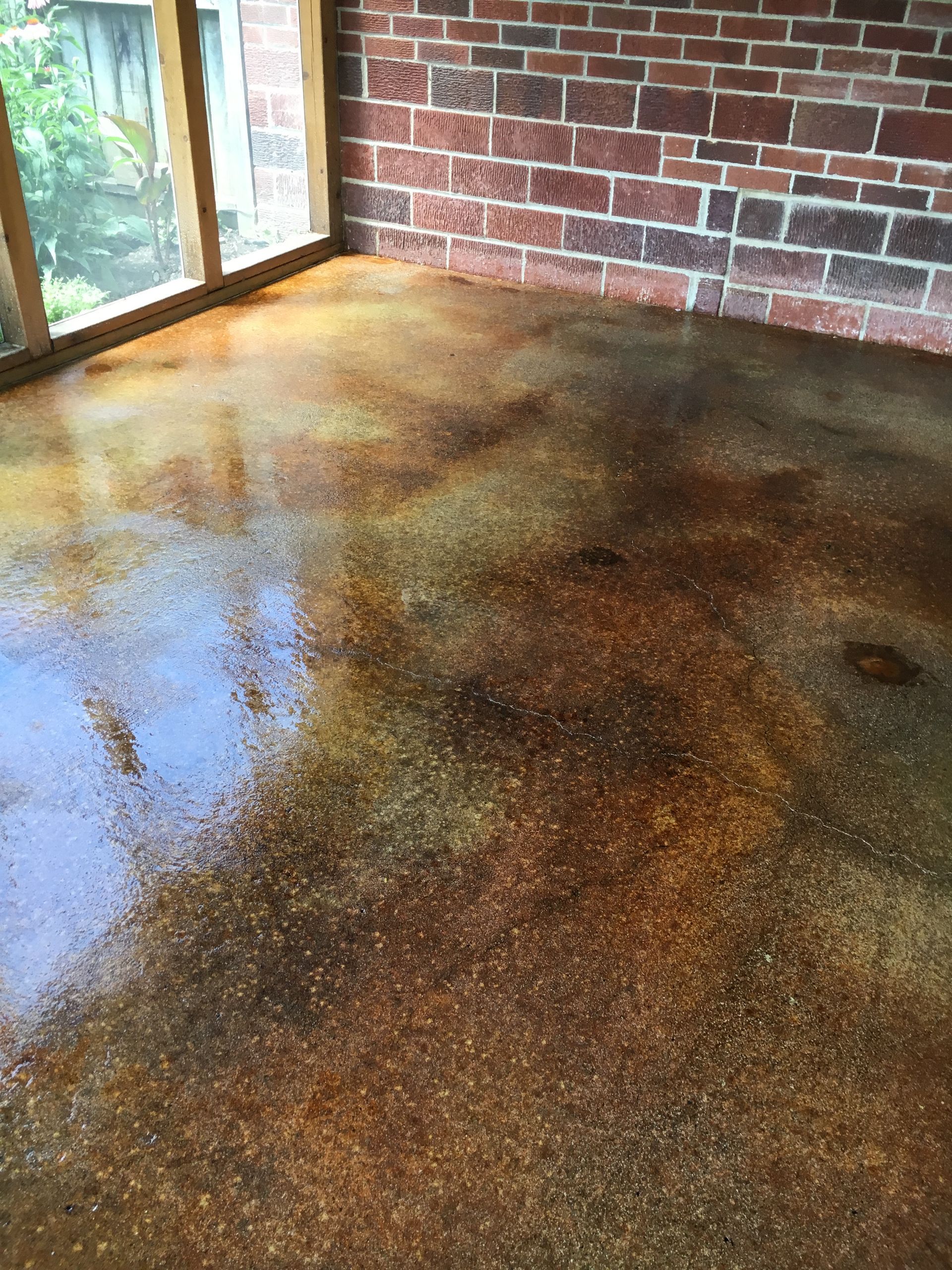 Stained concrete floor with brown and gold hues, by a brick wall and a window with sunlight.