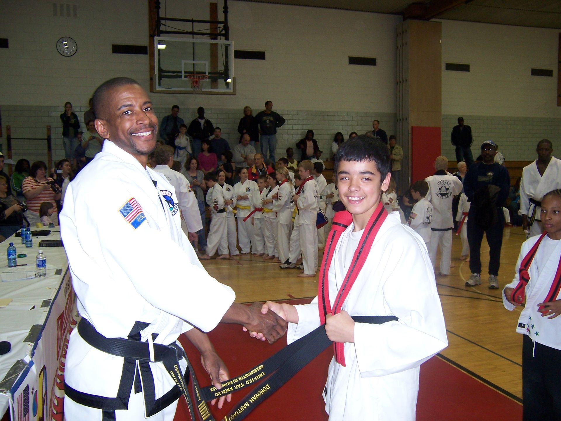 Two martial arts competitors shake hands at a gym ceremony, one wearing a medal and red belt.