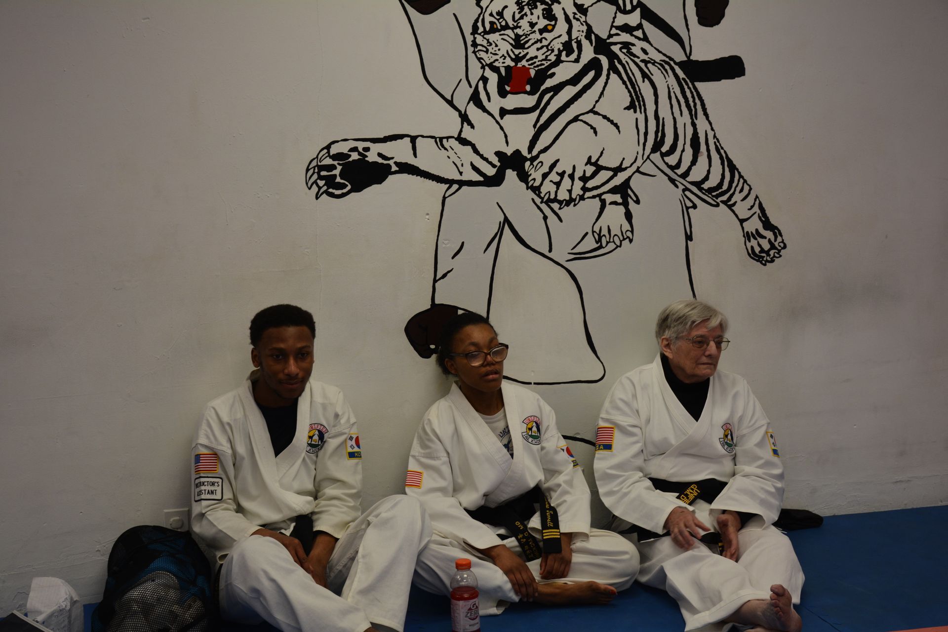 Three people in white martial arts uniforms sit on blue mats in a studio with a tiger mural on the wall.